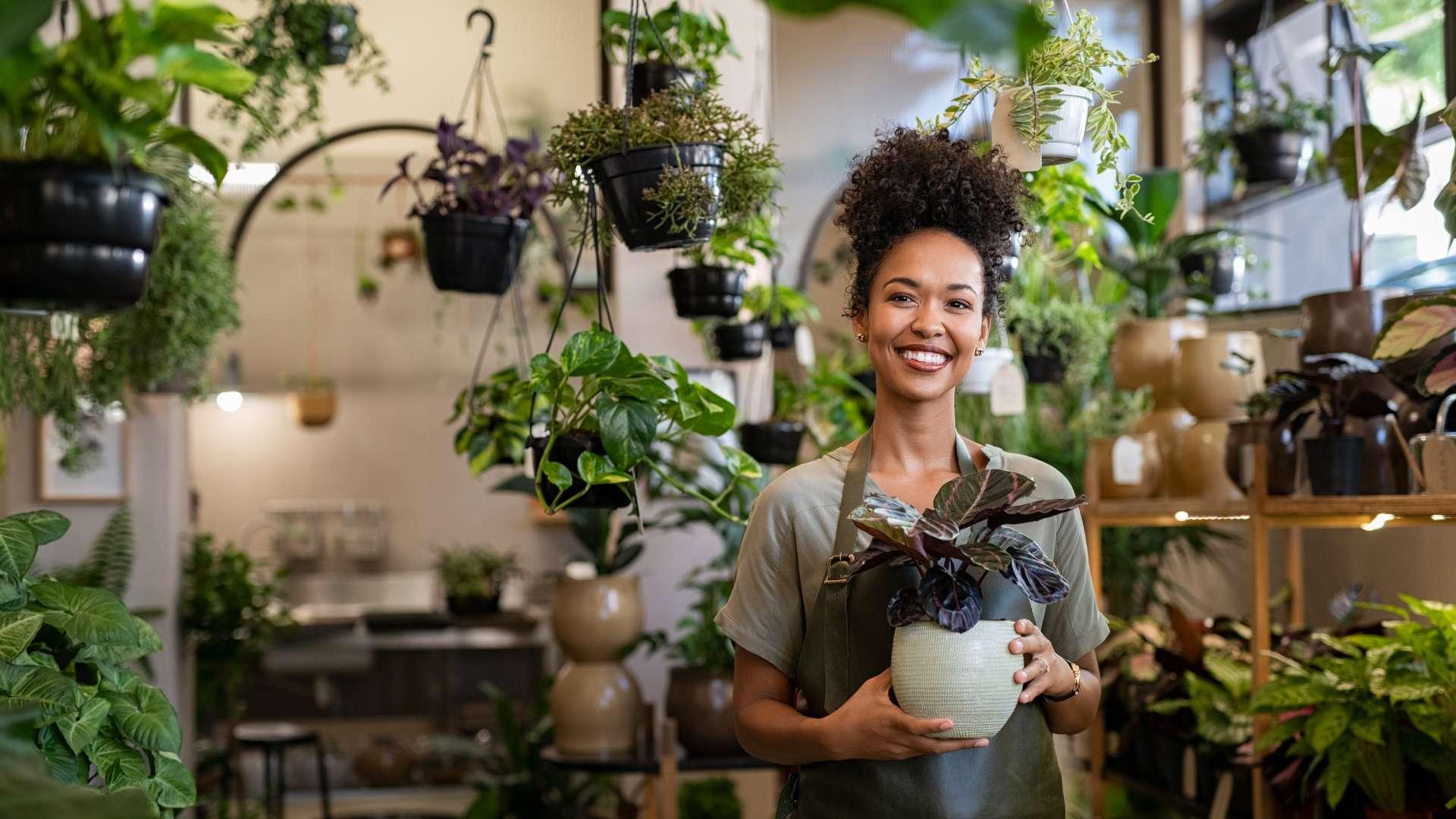 A smiling woman stands in her plant shop, surrounded by plants.